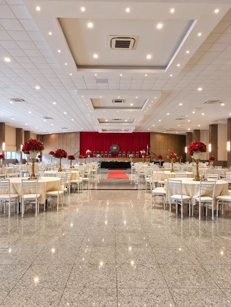 Salão montado pela Excellence: mesas redondas com toalha creme, cadeiras brancas e arranjos de rosas vermelhas em pedestais dourados